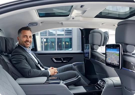A male passenger is sitting comfortably in the back seat of a luxury vehicle, smiling while using the in-car Wi-Fi shown on the screen, representing modern amenities and comfort.