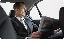 A close-up view of a male business passenger sitting in the back seat of a car, dressed in a suit and tie, looking outside as he reads a newspaper or magazine.