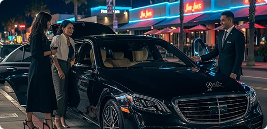 A chauffeur in a suit holds the door of a black Mercedes-Benz S-Class sedan open for two women on a vibrant city street at night.