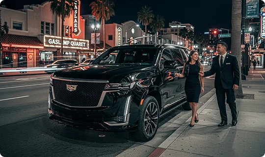 A chauffeur assists a woman in a black dress next to a black Cadillac Escalade SUV on a lively city street at night.