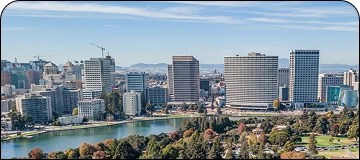 A panoramic view of a city skyline with modern high-rise buildings situated along a body of water (likely a lake or harbor) with a park in the foreground.