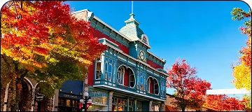 A vibrant, historic Victorian-style building with teal and red paint, featuring arched windows and a small tower, framed by trees with brilliant red and orange autumn foliage under a clear blue sky.