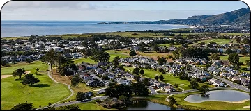 An aerial view of a coastal community featuring residential homes interspersed with green golf course fairways, a pond, and trees, all set against the blue ocean and distant rolling hills.