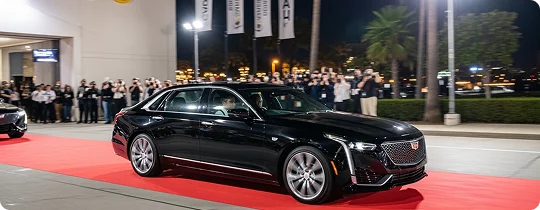A sleek black Cadillac CT6 drives on a red carpet at a glamorous event, surrounded by a cheering crowd and illuminated by spotlights against a nighttime cityscape with palm trees.