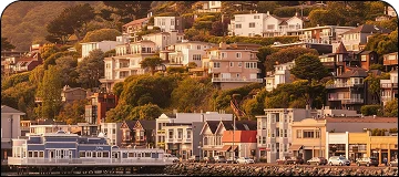 A view of the waterfront neighborhood of Sausalito, California, showing a row of colorful, commercial buildings and restaurants along the street level, including a prominent blue building on a pier over the water. Behind this strip, multi-story, residential homes are densely clustered up a steep, tree-covered hillside, illuminated by the warm light of sunset or sunrise.