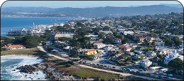 An elevated, panoramic view of a seaside town, likely Carmel-by-the-Sea or Monterey, California, showing densely packed homes and buildings along a rocky coastline, with a pier and buildings extending into a calm blue bay in the distance, backed by hazy mountains.