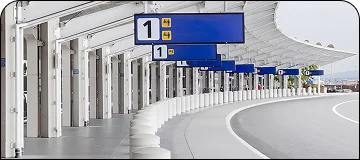 A bright white airport terminal drop-off lane with distant view of cars exiting it and blue sign boards visible.