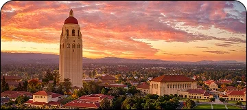 An elevated view of the Stanford University campus at sunset, featuring the prominent tall, light-colored Hoover Tower with a red-tiled roof, surrounded by other academic buildings and trees, all under a sky filled with dramatic orange and pink clouds.
