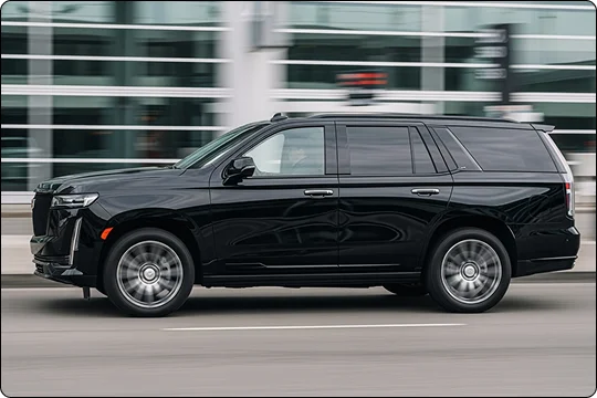 A sleek black SUV (Cadillac Escalade) is shown in a motion blur shot, driving quickly past a modern glass building.