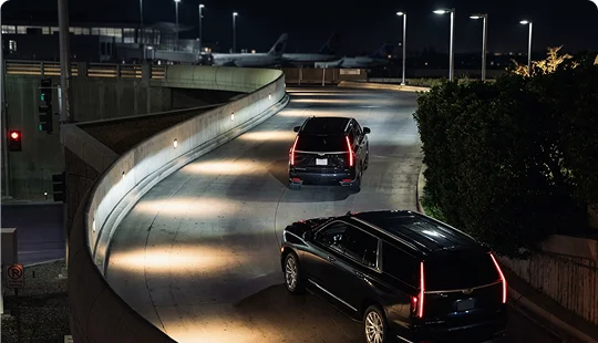 Two black luxury SUVs navigate a curved, illuminated terminal ramp at night. The elevated roadway overlooks an airport tarmac with commercial airplanes parked in the background.