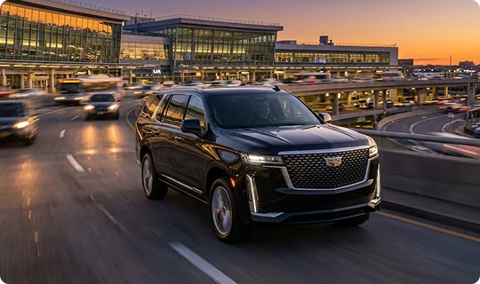 A black Cadillac Escalade driving on a highway ramp at sunset with an airport terminal in the background.