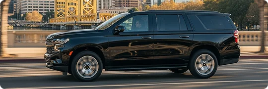 A black Chevrolet Tahoe SUV is driving on a city bridge or overpass. The motion blur suggests speed, and a prominent yellow-painted truss bridge and cityscape are visible in the background.
