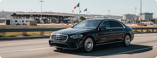 A black Mercedes-Benz S-Class sedan drives on a highway approaching a border crossing station with flags visible.