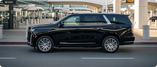 Side view of a Luxury Black SUV driven by a chauffeur in motion with blur wheels and background modern building visible in bright sunshine.