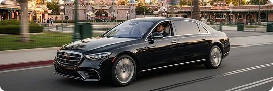 A black Mercedes-Benz S-Class luxury sedan is in motion on a road with a theme park entrance structure visible in the background, showing a chauffeur in a dark suit behind the wheel, suggesting a luxury travel experience to a resort or attraction.
