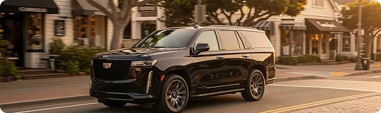 A sleek black Cadillac Escalade SUV drives through a sunny, charming downtown street lined with shops.