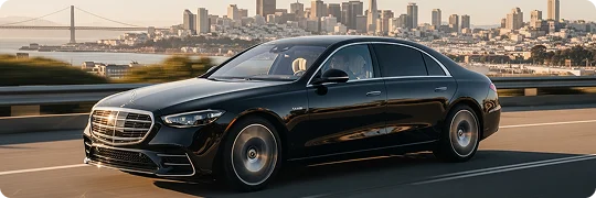 A black Mercedes-Benz S-Class sedan is driving on a sunlit freeway with the San Francisco skyline and the Bay Bridge visible in the background across the water. The image conveys movement, emphasizing the luxury vehicle and the iconic cityscape.