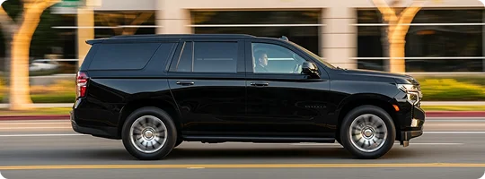 A side view of a black luxury SUV, likely a Chevrolet Suburban, being driven at speed on a city street with motion blur in the background.