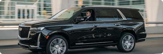 A black Cadillac Escalade SUV is driven by a chauffeur in a suit, seen from the side as it moves quickly past a modern glass and steel building (likely an airport or terminal). The wheels are blurred, suggesting motion.
