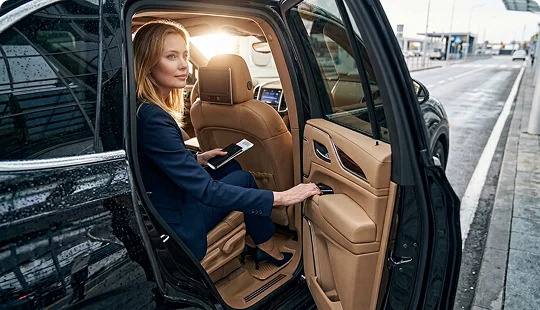 A businesswoman wearing a dark blazer sits in the tan leather backseat of a luxury SUV, looking outward with the door open. She holds a passport and a boarding pass in her hand, with an airport or transit road visible in the background.