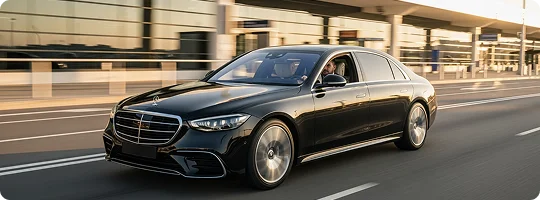 A black luxury sedan is driving quickly on a road next to an airport terminal during sunset. A person is visible in the driver's seat.