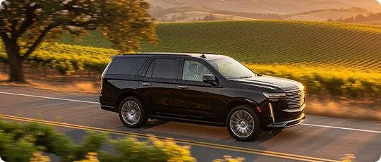 A luxurious black SUV (Cadillac Escalade) drives along a country road through sunny, rolling vineyard hills.