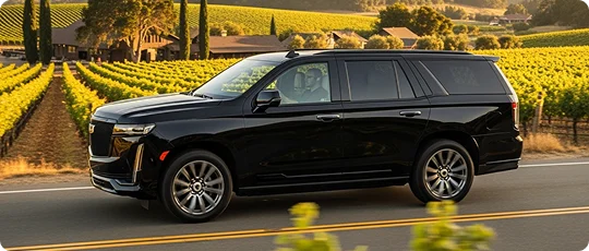 A side view of a black luxury SUV, likely a Cadillac Escalade, being driven on a road with rows of green grapevines and winery buildings in a vineyard setting on either side.