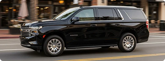 A black Chevrolet Tahoe SUV is driving quickly on a city street, creating a sense of motion blur in the background. The background features blurred storefronts and outdoor dining areas, suggesting a commercial or downtown setting.