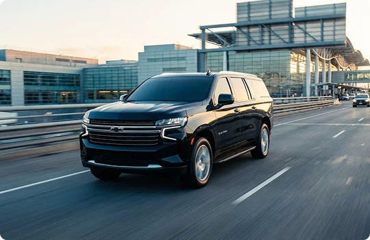 A black Chevrolet Suburban drives along a multi-lane highway toward a large, glass-walled airport terminal at dusk.