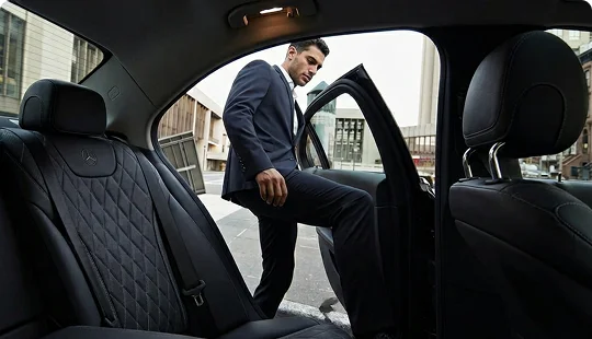 An interior view looking out of the open rear door of a luxury sedan with black quilted leather seats. A man in a dark business suit steps into the vehicle from a city street.