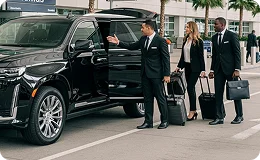 A suited chauffeur points at his polished new black Cadillac Escalade with its read door and trunk door opened while an elegant lady alongside a man is walking towards it carrying luggage on a palm lined street drop-off lane.