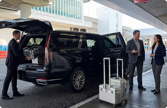 A chauffeur in a black suit loads a hard-case bag into the open trunk of a black luxury SUV. A man and a woman in business attire stand talking on the airport sidewalk next to two silver rolling suitcases.