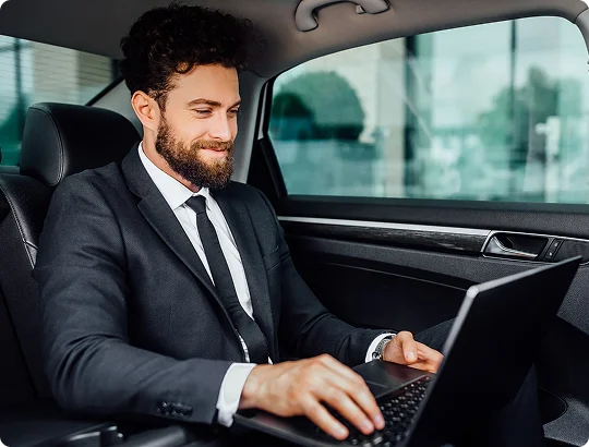 A businessman seated in the back of a premium car works on his laptop with a focused smile.