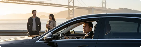 A chauffeur is seen in profile in the driver's seat of a black Mercedes-Benz sedan. Through the car's window, a couple stands by a guardrail, admiring the sunset view over a bay with a large suspension bridge visible in the distance.