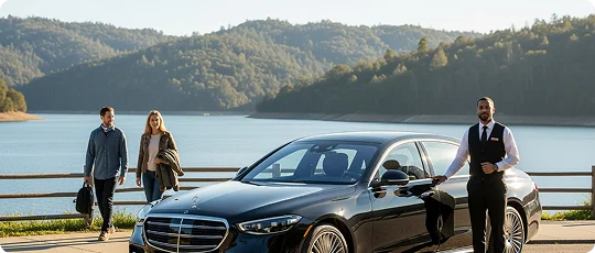 A couple walks by a waterfront with wooden grill behind them and a luxury black Mercedes Benz S Class is parked besides them with a suited chauffeur standing outside with it. Green forest mountains are visible in distant background at the other side of the lake or pond.