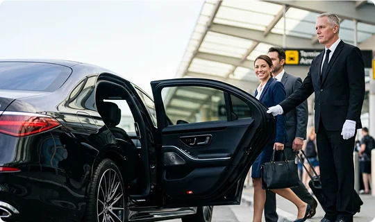 A professional chauffeur wearing a suit and white gloves holds open the back door of a black luxury sedan. A businesswoman and a man with luggage smile as they approach the vehicle outside an airport terminal.