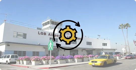 Exterior view of the Long Beach Airport terminal building with modern white facade, glass windows, palm trees, and parked cars in front under a bright blue sky.
