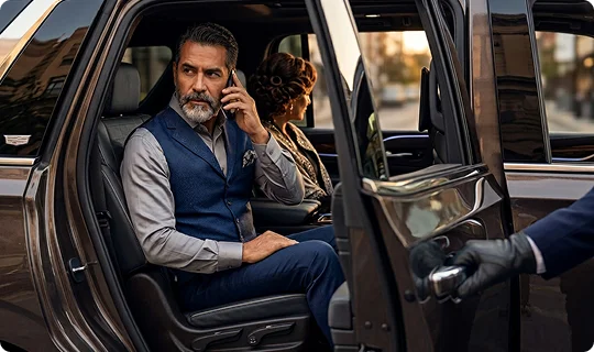 A man with a graying beard wearing a blue vest talks on a smartphone in the back seat of a luxury vehicle. A gloved hand holds the passenger door open while a woman sits beside him, looking out the window.