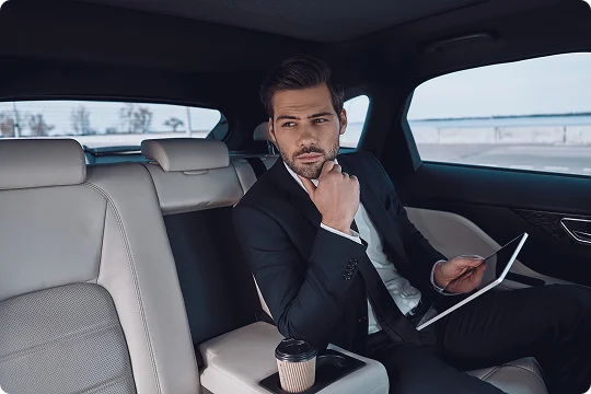 Businessman seated in a luxury car’s rear seat, holding a tablet with coffee beside him, gazing out thoughtfully.