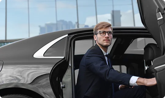 A professionally dressed man steps out of a sleek black luxury car, exuding business-class confidence near a modern glass-fronted terminal.
