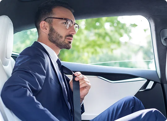 A sharply dressed businessman buckles up in the backseat of a luxury car, framed by sleek interior lines and lush greenery outside the window.