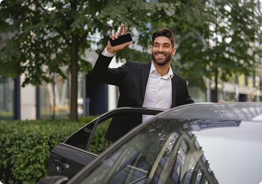 A cheerful man in a blazer steps out of a sleek black car, smiling and waving his phone in a lively, tree-lined urban setting.