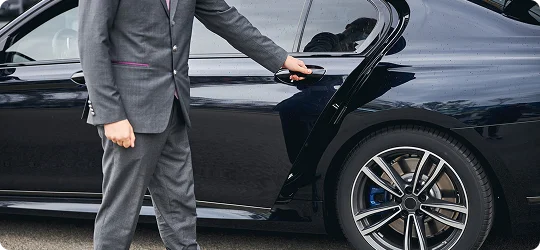 A chauffeur in a sharp gray suit opens the rear door of a luxury black sedan, ready to offer a refined, professional welcome.
