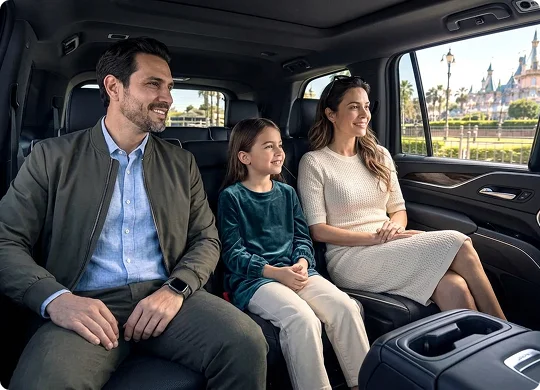 A family of three sits comfortably in the leather back seats of a luxury vehicle, smiling as they look out the window. Visible through the car window is an amusement park landscape featuring a large castle in the distance.