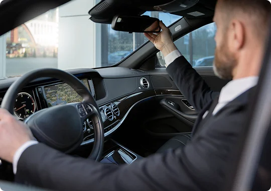An interior view of a luxury car's cockpit showing a bearded chauffeur in a suit and white shirt adjusting the rearview mirror with his right hand. The car's dashboard features a navigation screen and wood grain trim.