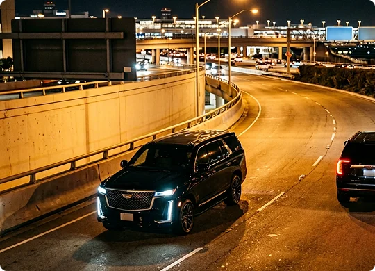 A black luxury SUV drives along a curved, multi-lane highway ramp at night with its headlights illuminated. The highway overhead is lit by streetlights, with city infrastructure and other vehicles visible in the background.