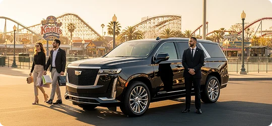 A chauffeur stands next to a black Cadillac Escalade as a couple walks in front of an amusement park.