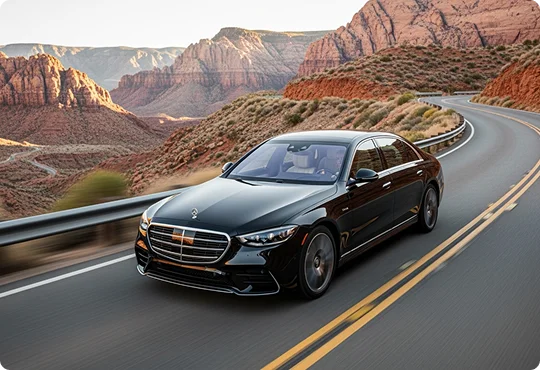 A black Mercedes S-Class sedan drives along a winding road through a picturesque desert canyon with red rock formations.