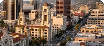 Broad evening view of Santa Ana's buildings and roadway with visible palm trees and cars parked on sideway in the center of this image.