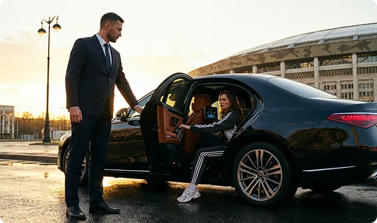  A formally dressed young couple just stepped out of a black luxury SUV from the rear seat and walks. A chauffeur holds the rear door open and watches them walk. Background shows a white SUB, a black sedan with two persons under a hotel entrance.